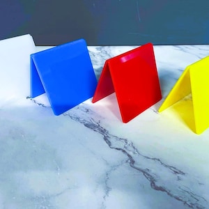 May include: Four colorful, folded acrylic signs in white, blue, red, and yellow. Each sign is folded in half, creating a triangular shape. The signs are arranged in a row on a marble-patterned surface, with a dark blue background.