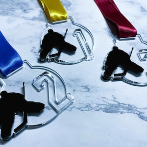 May include: Clear acrylic hockey medals with black player silhouettes and numbers 1, 2, and 3. Each medal has a different colored satin ribbon: blue, yellow, and red. The medals are displayed on a marble-patterned surface.