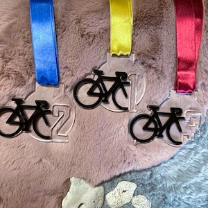 May include: Three clear acrylic medals with black bicycle silhouettes and numbers 2, 1, and 3. Each medal has a different colored ribbon: blue, yellow, and red. The medals are displayed on a textured surface.