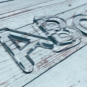 Könnte beinhalten: Klare Acrylbuchstaben, die das Wort "ABC" bilden, sind auf einer hellen Holzmaserung angeordnet. Die Buchstaben haben ein transparentes Aussehen mit dunklen Umrissen und erzeugen einen dreidimensionalen Effekt. Die Buchstaben sind abgewinkelt und werfen subtile Schatten.