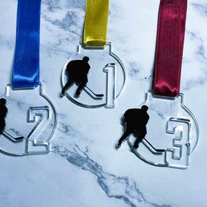 May include: Three clear acrylic hockey medals with blue, yellow, and burgundy ribbons. Each medal features a black silhouette of a hockey player and the numbers 1, 2, and 3. The medals are displayed on a marble-patterned surface.