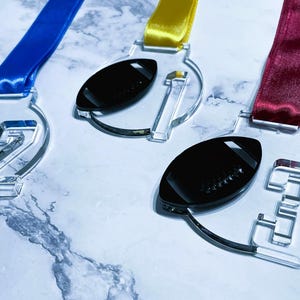 May include: Clear acrylic medals with black footballs and numbers 1, 2, and 3. Each medal has a different colored ribbon: blue, yellow, and burgundy. The medals are on a marble-patterned surface.