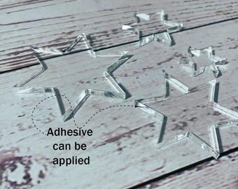 May include: Clear acrylic star shapes in various sizes. The text "Adhesive can be applied" is next to one of the stars. These stars are ideal for crafts and DIY projects. The stars are on a light wood grain surface.