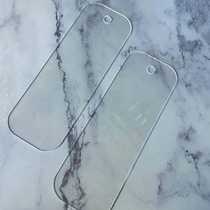 May include: Two clear, rectangular acrylic bookmarks with rounded corners and a small hole at the top for attaching a tassel or string. The bookmarks are transparent, allowing the underlying surface to be visible. The background is a marbled pattern.