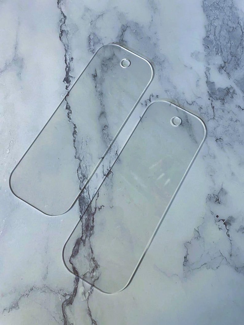 May include: Two clear, rectangular acrylic bookmarks with rounded corners and a small hole at the top for attaching a tassel or string. The bookmarks are transparent, allowing the underlying surface to be visible. The background is a marbled pattern.