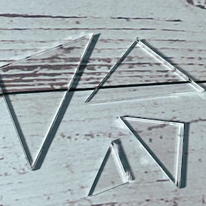 May include: Four clear, geometric shapes, including triangles and a quadrilateral, are arranged on a light-colored wooden surface. The shapes are transparent, casting shadows that add depth to the composition. The arrangement suggests a focus on geometry and design.