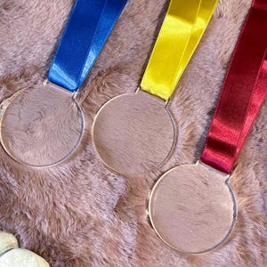 Può includere: Tre medaglie rotonde in acrilico trasparente con nastri blu, gialli e rossi. Le medaglie sono appese su uno sfondo strutturato marrone rosato. I nastri sono attaccati nella parte superiore di ogni medaglia.