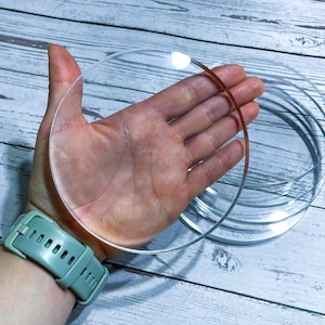 Puede incluir: Discos circulares de acrílico transparente de varios tamaños apilados y sostenidos en una mano. Se levanta un disco, mostrando su transparencia. Los discos están sobre una superficie clara con vetas de madera. Un reloj turquesa está en la muñeca.