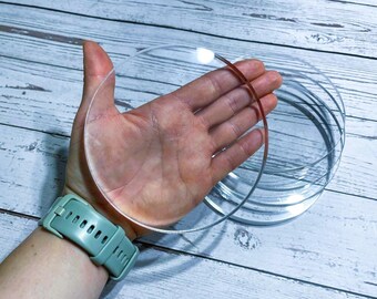 Puede incluir: Discos circulares de acrílico transparente de varios tamaños apilados y sostenidos en una mano. Se levanta un disco, mostrando su transparencia. Los discos están sobre una superficie clara con vetas de madera. Un reloj turquesa está en la muñeca.