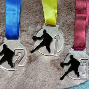 May include: Three clear acrylic basketball medals with black silhouettes of a player dribbling a ball. Each medal has a different number: 1, 2, and 3. The medals are suspended by blue, yellow, and red ribbons.