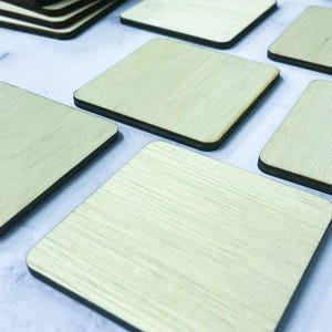 May include: A collection of square wooden coasters with a light beige wood grain finish and dark brown edges. The coasters are arranged in a scattered pattern, with one stack of several coasters visible in the upper left corner of the frame.