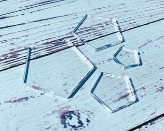 May include: Clear, geometric acrylic shapes in various sizes are arranged on a light blue wooden surface. The shapes include pentagons and hexagons, casting shadows. The objects are transparent and reflect light.