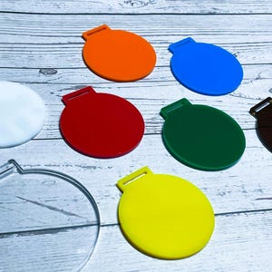 Può includere: Una collezione di ornamenti rotondi e colorati in vari colori, tra cui arancione, blu, rosso, verde, giallo, bianco e marrone. Ogni ornamento ha un piccolo anello nella parte superiore. Gli ornamenti sono disposti su una superficie chiara con venature del legno.