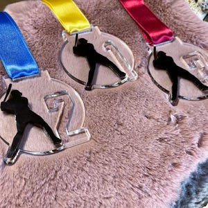 May include: Three clear acrylic baseball medals with colorful ribbons. Each medal features a black silhouette of a batter and a number. The ribbons are blue, yellow, and red. The medals are on a pink, furry surface.