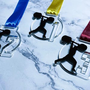 May include: Clear acrylic medals with black weightlifter silhouettes and numbers 1, 2, and 3. Each medal has a different colored ribbon: blue, yellow, and burgundy. The medals are on a marble-patterned surface.