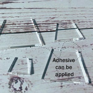 Pode incluir: Formas de acrílico transparente em vários tamanhos, incluindo trapézios e um quadrado menor. O texto "Adhesive can be applied" é visível, sugerindo projetos de artesanato ou DIY.