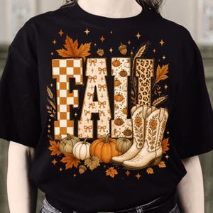 May include: A black t-shirt featuring the word FALL in large block letters with varied patterns including orange checkers, bows, pumpkins, and leopard print. The design includes orange autumn leaves, wheat stalks, pumpkins, and two cowboy boots.