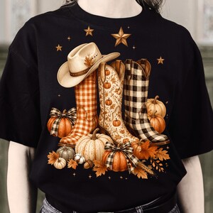 Puede incluir: Una camiseta negra con un estampado gráfico de tres botas vaqueras, una con cuadros naranjas, otra con un patrón de calabazas y hojas, y otra con cuadros negros. Un sombrero vaquero beige descansa encima, rodeado de calabazas y hojas de otoño.