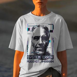 May include: White t-shirt featuring a grayscale portrait with glitch effects. The text "SYSTEM ERROR MUGSHOT_#400004" is printed below the portrait. The shirt is worn by a person with short, light-colored hair.