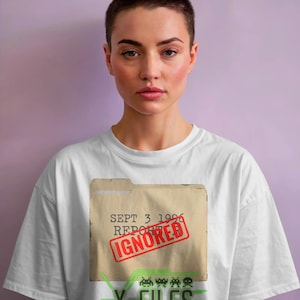 Puede incluir: Camiseta blanca con un diseño de Expediente X. El diseño incluye una carpeta con el texto "SEPT 3 1996 REPORTED IGNORED" y las palabras "X-FILES THE TRUTH IS OUT THERE" en verde.