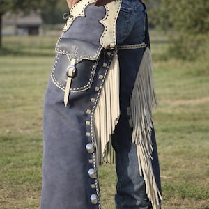 May include: Blue suede leather chaps with cream-colored accents, fringe, and silver-tone button details. The chaps feature a pocket and are worn over blue jeans. A classic Western wear item.