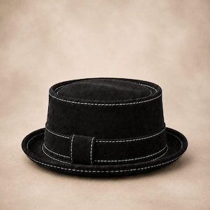 May include: A black pork pie hat featuring a flat crown and a short, upturned brim. The hat is constructed from a matte, textured material and is accented with prominent white contrast stitching along the crown, the brim, and the hat band.