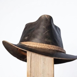 May include: A distressed brown leather hat with a woven band. The hat has a wide brim and a slightly curved crown. The hat is displayed on a wooden post against a white background.