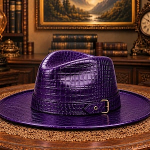 May include: A deep purple fedora hat featuring a glossy, embossed crocodile skin texture. The hat includes a matching purple band around the crown, secured with a gold-toned metal buckle on the side, and a wide, flat brim.
