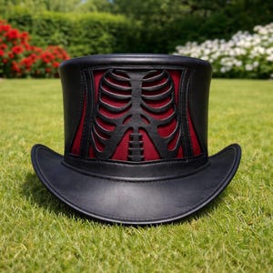 May include: Black top hat with a red interior and a cut-out rib cage design. The hat has a wide brim and is made of leather. The hat is sitting on green grass with red and white flowers in the background.