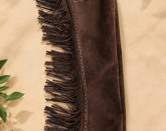May include: A dark brown suede leather chap featuring long fringe along the outer leg. The upper portion includes a decorative embossed floral pattern on a darker leather yoke with a silver-toned metal buckle and a thin adjustable strap.