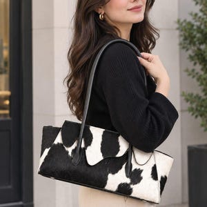 Könnte beinhalten: Eine rechteckige Handtasche mit schwarz-weißem Kuhfellmuster und einer strukturierten Oberfläche. Die Tasche verfügt über einen schwarzen Ledergriff und einen Klappenverschluss mit einem wellenförmigen Rand entlang der oberen Öffnung.