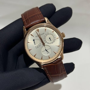 Puede incluir: Un reloj de pulsera dorado con esfera clara y correa de cuero marrón. La esfera del reloj presenta múltiples subesferas y el texto "Jaeger-LeCoultre". La correa es de color marrón oscuro, y el reloj tiene un diseño clásico y elegante.