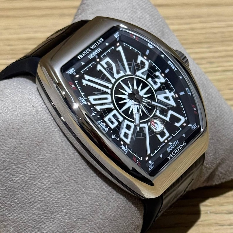 May include: A luxury Franck Muller watch with a black dial and white numerals. The watch has a silver-tone case and a black leather strap. The dial features a compass rose design and the words "SOUTH YACHTING".