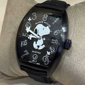 Könnte beinhalten: Schwarze Armbanduhr mit schwarzem Armband. Das Zifferblatt zeigt eine weiße Snoopy-Grafik, die einen Knochen hält, mit den Worten "FRANCK MULLER" und "BAMFORD" auf dem Zifferblatt. Die Uhr hat ein schwarzes Gehäuse und eine blaue Krone.