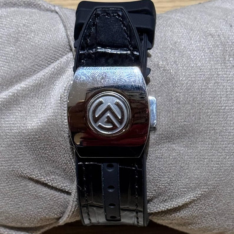May include: Close-up of a black watch band with a silver buckle. The buckle features a circular emblem with a stylized design. The band appears to be made of leather and rubber.
