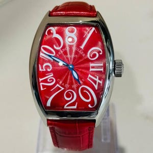 Op de afbeelding: Een luxe polshorloge met een rode wijzerplaat en een rode leren band. De wijzerplaat van het horloge heeft grote witte cijfers en de woorden "FRANCK MULLER GENEVE CRAZY HOURS". Het horloge heeft een zilverkleurige kast.