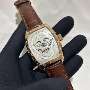 May include: A luxury wristwatch with a brown leather strap and a gold-toned case. The watch face features a detailed skull design with the words "FRANCK MULLER GENEVE" and "SWISS MADE". The watch is held by a gloved hand.