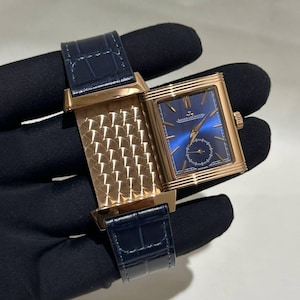 Peut inclure: Une montre rectangulaire en or rose avec un cadran bleu et un bracelet en cuir bleu foncé. Le cadran de la montre comporte un petit cadran des secondes et la marque "Jaeger-LeCoultre". La montre est ouverte, révélant un intérieur doré à motifs.