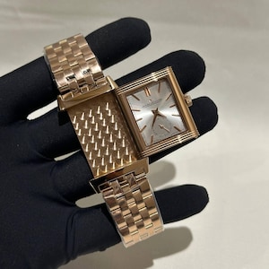 May include: A rose gold rectangular wristwatch with a silver dial and a small seconds subdial. The watch has a matching rose gold bracelet. The watch is open, revealing the intricate design of the back.