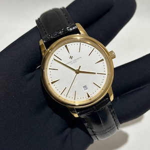 May include: A gold wristwatch with a white face, gold hands and markers, and a black leather strap. The watch has a date display at the 3 o'clock position. The watch face displays the text "VACHERON CONSTANTIN GENEVE".