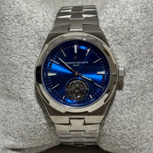 Pode incluir: Um relógio de pulso em aço inoxidável com um mostrador azul vibrante. O relógio apresenta um mecanismo complexo visível através do mostrador, com marcadores de hora e ponteiros brancos. A pulseira é feita de aço inoxidável. O relógio está sobre uma superfície cinza.
