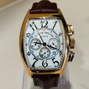 May include: A gold-toned wristwatch with a white face and black numerals. The watch features multiple sub-dials, a brown leather strap, and the text "FRANCK MULLER GENEVE SWISS MADE".