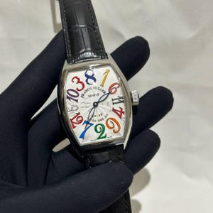 Puede incluir: Un reloj de pulsera plateado con esfera blanca y correa de cuero negro. La esfera del reloj presenta números coloridos y las palabras "CRAZY OR DREAMS". El reloj tiene forma rectangular y una corona plateada.