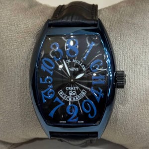 May include: A black and blue Franck Muller Crazy Hours Black Badge watch with a black leather band. The watch face features blue numerals and the words "Crazy Black Badge" and "Neve". The watch is marked "Swiss Made".