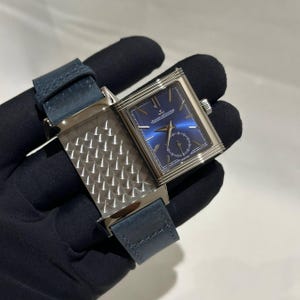 Puede incluir: Un reloj de pulsera rectangular con esfera azul y caja plateada. El reloj presenta un pequeño subdial de segundos y una correa de cuero azul. El reloj se sostiene sobre un fondo negro.