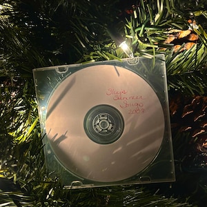 May include: A clear plastic CD jewel case containing a silver disc with handwritten red ink text that reads Sky's Summer Songs 2008. The case is positioned among green artificial pine needles and small glowing white holiday lights.