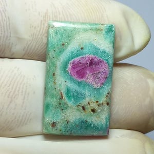 Könnte beinhalten: Ein rechteckiger Edelstein mit einer lebendigen Farbmischung. Der Stein weist einen zentralen, auffälligen fuchsiafarbenen Einschluss auf, der vor einem Hintergrund aus wirbelnden Türkis- und Grüntönen liegt. Kleine braune Flecken verleihen der Oberfläche des Steins Textur.