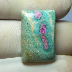 Könnte beinhalten: Ein rechteckiger Edelstein mit türkisfarbener und beigefarbener Grundfarbe, der lebendige rosa Einschlüsse aufweist. Der Stein wird von einer behandschuhten Hand gehalten, die seine polierte Oberfläche und natürlichen Muster zeigt. Dieser Edelstein ist ideal für die Schmuckherstellung.