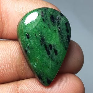 Könnte beinhalten: Ein polierter, tropfenförmiger Edelstein. Der Stein ist ein leuchtendes Grün mit schwarzen Einschlüssen, die ein gesprenkeltes Muster erzeugen. Der Edelstein ist glatt und reflektiert das Licht, wodurch seine natürliche Schönheit zur Geltung kommt. Die Form ist symmetrisch.