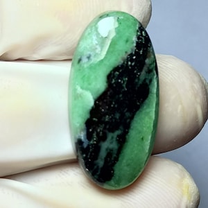 May include: Oval-shaped gemstone with a polished surface. The stone features a vibrant green base color with a striking black pattern running through the center. White and lighter green areas add contrast. The gemstone is held by a gloved hand.